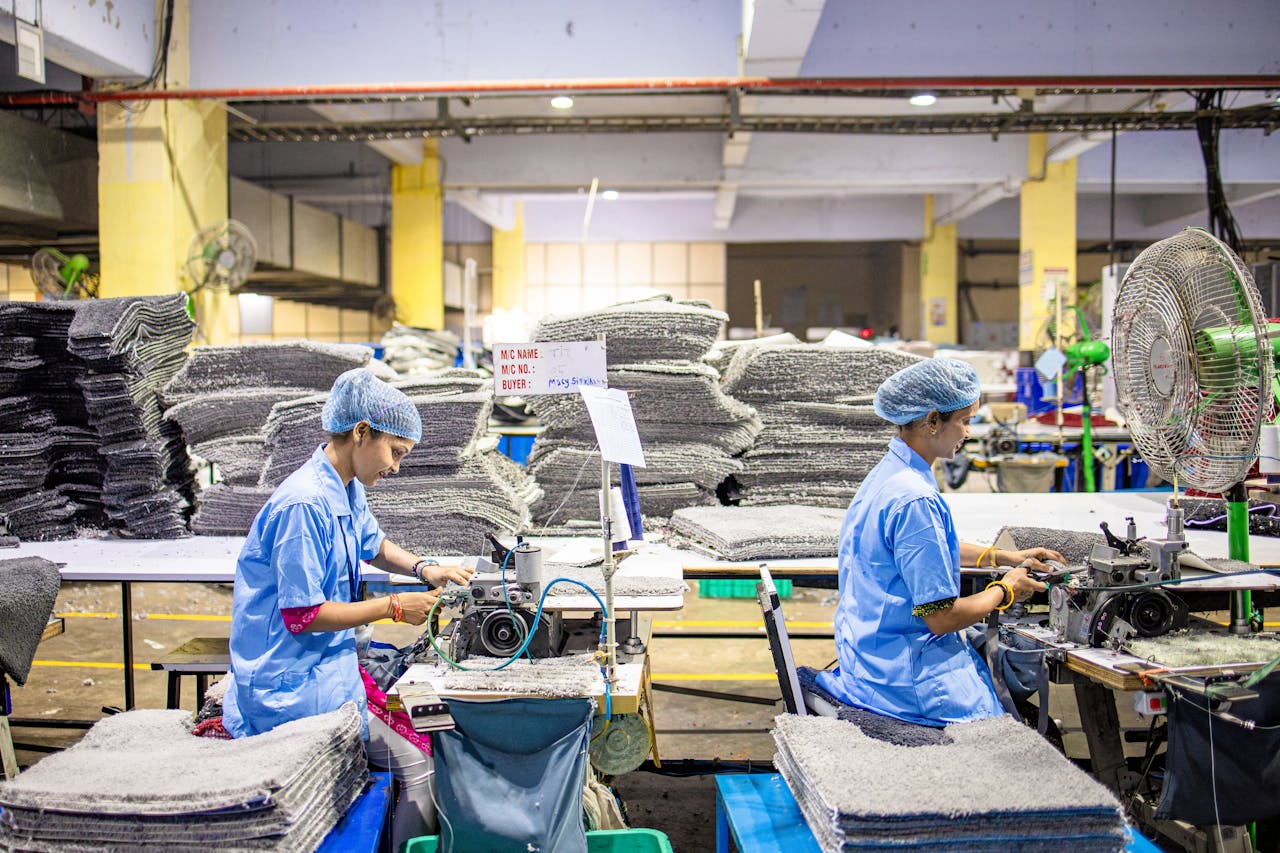 Two female textile workers operating sewing machines in a busy factory setting.